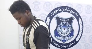 شخص مضبوط من جانب وحدة التحري يقف أمام شعار مديرية أمن تاجوراء بمركز الشرطة.