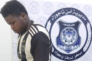 شخص مضبوط من جانب وحدة التحري يقف أمام شعار مديرية أمن تاجوراء بمركز الشرطة.