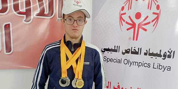 السباح عبد الهادي الرطيب يرتدي عدة ميداليات رياضية أمام لوحة تحمل شعار الأولمبياد الخاص الليبي.