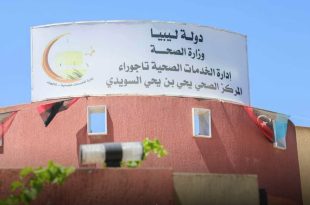 صورة لافتة بيضاء كبيرة تعتلي مبنى المركز الصحي يحيى بن يحيى السويدي في تاجوراء، مكتوب عليها "دولة ليبيا - وزارة الصحة - إدارة الخدمات الصحية تاجوراء - المركز الصحي يحيى بن يحيى السويدي"، ويظهر على جانبها شعار إدارة الخدمات الصحية، بينما تتدلى من المبنى أعلام ليبيا الصغيرة تحت سماء زرقاء صافية