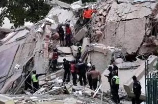 لقطة حية تُظهر مبنى سكنيًا منهارًا بالكامل ومتحولًا إلى ركام من الخرسانة والأعمدة المحطمة جراء زلزال فنزويلا، ويتواجد في الموقع عدد كبير من أفراد فرق الإنقاذ والشرطة الذين يرتدون خوذًا وسترات واقية أثناء بحثهم بين الأنقاض عن ناجين.