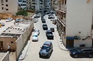 لقطة علوية تظهر رتلاً طويلًا من سيارات الدفع الرباعي والمدرعات السوداء والرمادية التابعة لإدارة إنفاذ القانون وهي تتحرك داخل شارع النصر السكني في طرابلس، وتحيط بها عمارات سكنية وسيارات مدنية متوقفة على جانبي الطريق