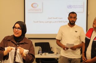 ورشة عمل تدريبية؛ تظهر في مقدمة الصورة جهة اليسار سيدة ترتدي حجاباً أسود ونظارة طبية وقميصاً بنياً وتتحدث بحماس موجهةً إشارات بيديها. وفي الخلفية جهة اليمين يقف رجلان، أحدهما يرتدي قميصاً أبيض ويمسك بدفتر صغير وقلم، والآخر يبتسم ويرتدي سترة (فيست) باللونين الأحمر والأسود تحمل شعار المركز الوطني لمكافحة الأمراض