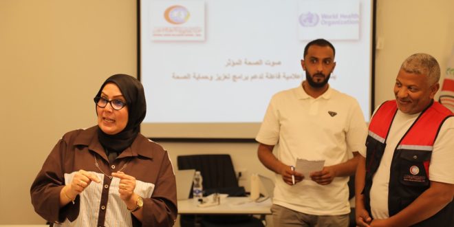 ورشة عمل تدريبية؛ تظهر في مقدمة الصورة جهة اليسار سيدة ترتدي حجاباً أسود ونظارة طبية وقميصاً بنياً وتتحدث بحماس موجهةً إشارات بيديها. وفي الخلفية جهة اليمين يقف رجلان، أحدهما يرتدي قميصاً أبيض ويمسك بدفتر صغير وقلم، والآخر يبتسم ويرتدي سترة (فيست) باللونين الأحمر والأسود تحمل شعار المركز الوطني لمكافحة الأمراض
