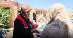 امرأة ترتدي سترة حمراء وسوداء وحجاباً تقدم مطويات تلوين وإرشادات صحية لامرأة أخرى ترتدي حجاباً بيج، ويظهر في الخلفية رجل يحمل مطويات وامرأة أخرى، مع لقطة خارجية أمام مبنى صحي في يوم مشمس.