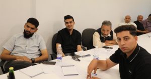 لقطة أفقية من داخل قاعة التدريب تُظهر مجموعة من الرجال والإعلاميين يجلسون حول طاولة اجتماعات بيضاء طويلة. يظهر في مقدمة الصورة جهة اليمين شاب يرتدي قميصاً أسود يلتفت نحو الكاميرا ويمسك بقلم، ويجلس خلفه على امتداد الطاولة عدة مشاركين؛ أحدهم يرتدي قميصاً أسود ويبتسم، وآخر ملتحٍ يرتدي قميصاً رمادياً فاتحاً مخططاً، ورجل أكبر سناً يرتدي صديرياً أسود ويضع يديه على الطاولة، بالإضافة إلى مشاركين آخرين