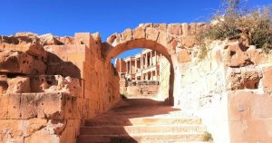 ممر حجري قديم تعلوه قنطرة (قوس) أثرية مبنية من الحجر الرملي في مدينة صبراتة، يظهر من خلاله جزء من أعمدة المسرح الروماني الشهير تحت سماء زرقاء صافية