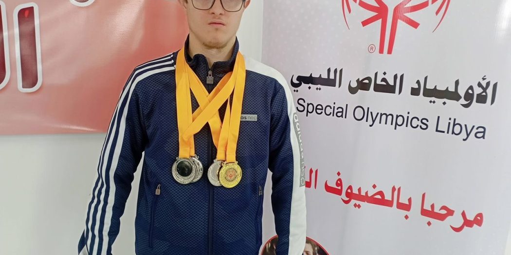 السباح عبد الهادي الرعيض يرتدي عدة ميداليات رياضية أمام لوحة تحمل شعار الأولمبياد الخاص الليبي.