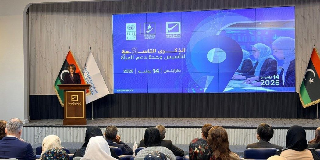 قاعة مؤتمرات تضم حضورًا من الرجال والنساء يجلسون أمام منصة تتحدث عليها امرأة، وخلفها شاشة كبيرة تعرض شعار الذكرى التاسعة لتأسيس وحدة دعم المرأة في طرابلس بتاريخ 14 يونيو 2026، إلى جانب أعلام ليبيا وشعارات الجهات المنظمة.