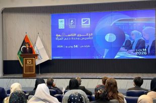 قاعة مؤتمرات تضم حضورًا من الرجال والنساء يجلسون أمام منصة تتحدث عليها امرأة، وخلفها شاشة كبيرة تعرض شعار الذكرى التاسعة لتأسيس وحدة دعم المرأة في طرابلس بتاريخ 14 يونيو 2026، إلى جانب أعلام ليبيا وشعارات الجهات المنظمة.
