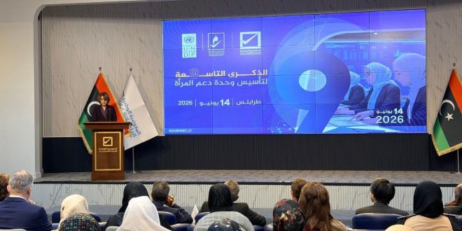 قاعة مؤتمرات تضم حضورًا من الرجال والنساء يجلسون أمام منصة تتحدث عليها امرأة، وخلفها شاشة كبيرة تعرض شعار الذكرى التاسعة لتأسيس وحدة دعم المرأة في طرابلس بتاريخ 14 يونيو 2026، إلى جانب أعلام ليبيا وشعارات الجهات المنظمة.