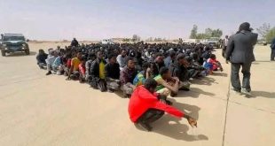 صورة تعبيرية تُظهر حشدًا كبيرًا من المهاجرين وطالبي اللجوء محتجزين على أرضية خرسانية مكشوفة في العراء داخل ما يبدو أنه مركز احتجاز في ليبيا. يجلس مئات الرجال جنبًا إلى جنب في صفوف طويلة، وأغلبهم ينظر لأسفل أو بعيدًا عن الكاميرا. في المقدمة، يظهر مهاجر يرتدي قميصًا أحمر ساطعًا وهو يجلس القرفصاء. على يسار الصورة، توجد سيارة دفع رباعي مصفحة (تويوتا) بيضاء وسوداء تابعة على الأرجح للأجهزة الأمنية الليبية، بينما يقف عدة رجال في ملابس مدنية وسترات رسمية بالقرب من المهاجرين المحتجزين على اليمين، أحدهم يصور المشهد. الخلفية عبارة عن منطقة صحراوية مسطحة مع بعض الأشجار والمباني البسيطة البعيدة تحت سماء ضبابية.