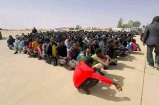 صورة تعبيرية تُظهر حشدًا كبيرًا من المهاجرين وطالبي اللجوء محتجزين على أرضية خرسانية مكشوفة في العراء داخل ما يبدو أنه مركز احتجاز في ليبيا. يجلس مئات الرجال جنبًا إلى جنب في صفوف طويلة، وأغلبهم ينظر لأسفل أو بعيدًا عن الكاميرا. في المقدمة، يظهر مهاجر يرتدي قميصًا أحمر ساطعًا وهو يجلس القرفصاء. على يسار الصورة، توجد سيارة دفع رباعي مصفحة (تويوتا) بيضاء وسوداء تابعة على الأرجح للأجهزة الأمنية الليبية، بينما يقف عدة رجال في ملابس مدنية وسترات رسمية بالقرب من المهاجرين المحتجزين على اليمين، أحدهم يصور المشهد. الخلفية عبارة عن منطقة صحراوية مسطحة مع بعض الأشجار والمباني البسيطة البعيدة تحت سماء ضبابية.