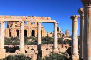 صفوف من الأعمدة الرومانية القديمة ذات التيجان المنحوتة المقامة على قواعد حجرية بمدينة صبراتة، يظهر في خلفيتها مبنى المسرح الأثري ذو الأقواس تحت سماء زرقاء صافية وتتخللها شجيرات خضراء