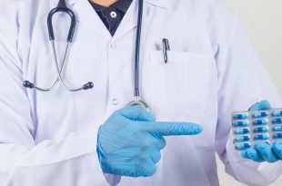 طبيب يرتدي معطفاً أبيض وقفازات طبية زرقاء ويشير بيده إلى شريط من كبسولات المضاد الحيوي.