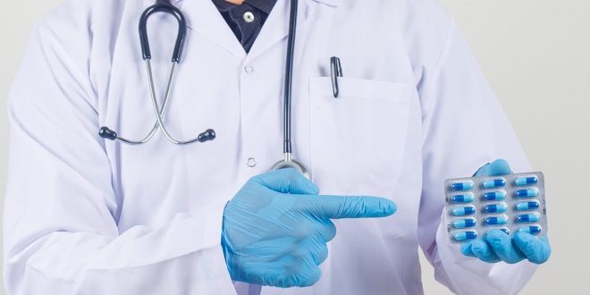 طبيب يرتدي معطفاً أبيض وقفازات طبية زرقاء ويشير بيده إلى شريط من كبسولات المضاد الحيوي.