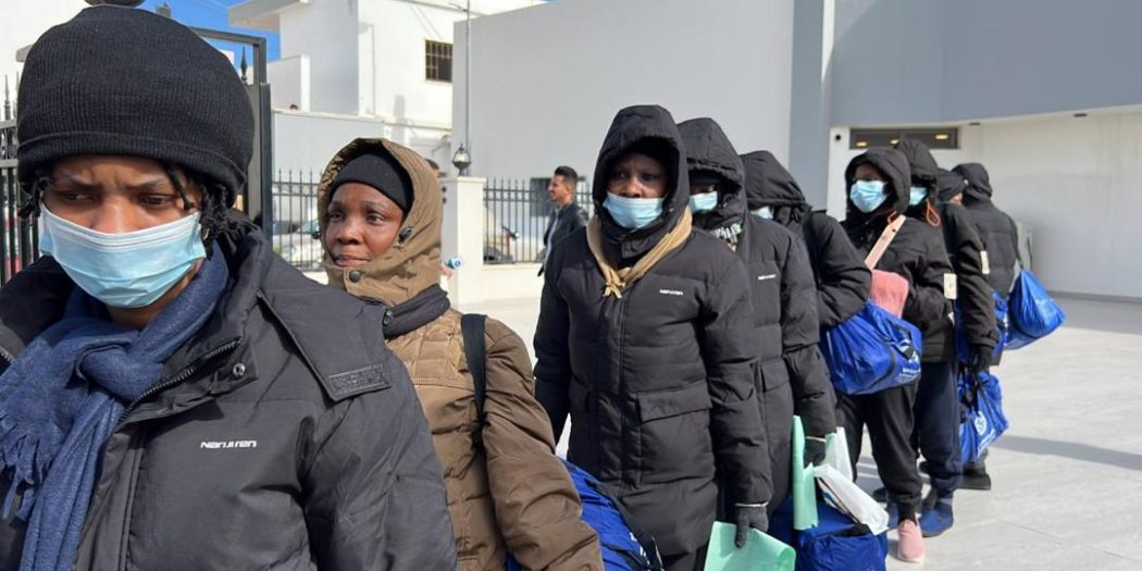 مجموعة من اللاجئين والمهاجرين يصطفون في ممر خارجي مرتدين كمامات ومعاطف شتوية ويحملون حقائب زرقاء أثناء إجراءات إجلاء أو إغاثة