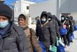 مجموعة من اللاجئين والمهاجرين يصطفون في ممر خارجي مرتدين كمامات ومعاطف شتوية ويحملون حقائب زرقاء أثناء إجراءات إجلاء أو إغاثة