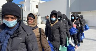مجموعة من اللاجئين والمهاجرين يصطفون في ممر خارجي مرتدين كمامات ومعاطف شتوية ويحملون حقائب زرقاء أثناء إجراءات إجلاء أو إغاثة