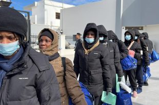 مجموعة من اللاجئين والمهاجرين يصطفون في ممر خارجي مرتدين كمامات ومعاطف شتوية ويحملون حقائب زرقاء أثناء إجراءات إجلاء أو إغاثة