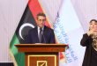 رئيس المفوضية الوطنية العليا للانتخابات عماد السايح يلقي كلمة خلف منصة خشبية، وبجانبه علم ليبيا وشعار المفوضية، وإلى يمينه مترجمة لغة الإشارة أثناء الاحتفالية السنوية لوحدة دعم المرأة.
