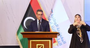رئيس المفوضية الوطنية العليا للانتخابات عماد السايح يلقي كلمة خلف منصة خشبية، وبجانبه علم ليبيا وشعار المفوضية، وإلى يمينه مترجمة لغة الإشارة أثناء الاحتفالية السنوية لوحدة دعم المرأة.