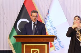 رئيس المفوضية الوطنية العليا للانتخابات عماد السايح يلقي كلمة خلف منصة خشبية، وبجانبه علم ليبيا وشعار المفوضية، وإلى يمينه مترجمة لغة الإشارة أثناء الاحتفالية السنوية لوحدة دعم المرأة.