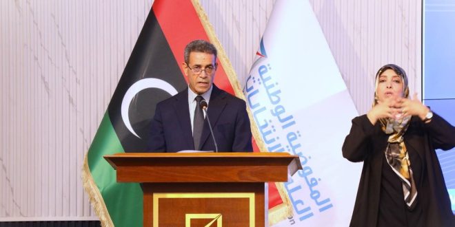 رئيس المفوضية الوطنية العليا للانتخابات عماد السايح يلقي كلمة خلف منصة خشبية، وبجانبه علم ليبيا وشعار المفوضية، وإلى يمينه مترجمة لغة الإشارة أثناء الاحتفالية السنوية لوحدة دعم المرأة.