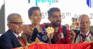 ​لقطة احتفالية لبعثة المنتخب الليبي المدرسي للموي تاي تُظهر أحد الأبطال المتوجين يحمل ميدالية ذهبية وإلى جانبه أعضاء من الطاقم الفني والإداري يحملون العلم الليبي في بطولة العالم للمدارس بماليزيا 2026.