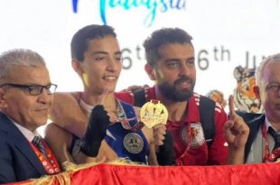 ​لقطة احتفالية لبعثة المنتخب الليبي المدرسي للموي تاي تُظهر أحد الأبطال المتوجين يحمل ميدالية ذهبية وإلى جانبه أعضاء من الطاقم الفني والإداري يحملون العلم الليبي في بطولة العالم للمدارس بماليزيا 2026.