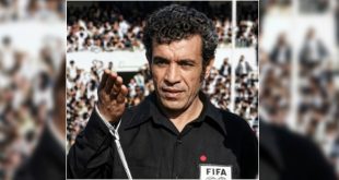 لقطة مقربة للحكم الدولي الليبي الراحل يوسف الغول بزي التحكيم الأسود الشعار الدولي "FIFA"، يرفع يده أثناء إدارة مباراة وسط حضور جماهيري.