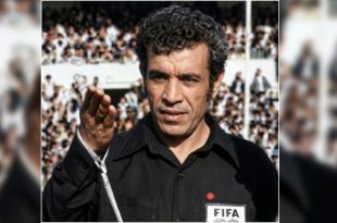 لقطة مقربة للحكم الدولي الليبي الراحل يوسف الغول بزي التحكيم الأسود الشعار الدولي "FIFA"، يرفع يده أثناء إدارة مباراة وسط حضور جماهيري.