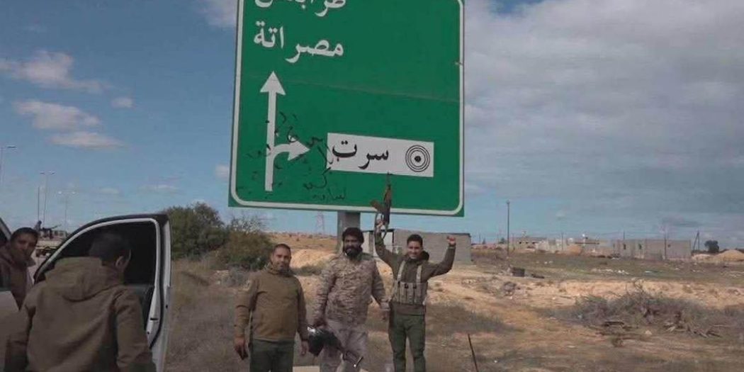 "عدة أشخاص يقفون بجوار لافتة طرق خضراء تشير إلى طرابلس ومصراتة وسرت، بالقرب من طريق في منطقة مفتوحة ذات طبيعة صحراوية"