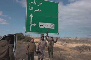 "عدة أشخاص يقفون بجوار لافتة طرق خضراء تشير إلى طرابلس ومصراتة وسرت، بالقرب من طريق في منطقة مفتوحة ذات طبيعة صحراوية"