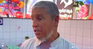 لقطة جانية للمواطن الحاج محمد الهادي، رجل مسن ذو شعر ولحية بيضاء يرتدي جلباباً فاتح اللون، يتحدث من داخل سوق أسماك محلي وتظهر خلفه لافتة تجارية وجدار سيراميك أبيض مع شعار منصة الصباح.