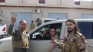 مجموعة من الرجال يقفون معًا في ساحة خارجية ويرفع العديد منهم علامة النصر بأيديهم، بينما تظهر مركبات وآليات عسكرية في الخلفية.