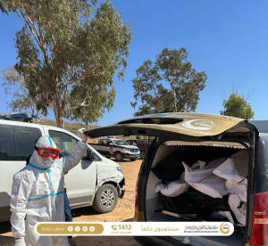 لقطة من مركز طب الطوارئ والدعم تظهر أحد أفراد الطاقم بملابس وقائية كاملة بجانب الصندوق الخلفي المفتوح لسيارة إسعاف ممتلئة بأكياس جثامين بيضاء وسوداء، في منطقة مفتوحة بها أشجار تحت سماء صافية.