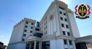 لقطة من زاوية منخفضة تُظهر مبنى مكتب النائب العام في طرابلس ذو التصميم المعماري الحديث واللون الأبيض تحت سماء صافية، مع وجود شعار النيابة العامة الليبية مدمجاً في أعلى يمين الصورة.