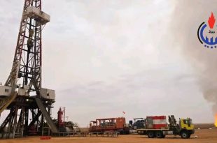 برج حفر ونفط ضخم تابع لشركة سرت لإنتاج وتصنيع النفط والغاز في حقل متخندوش بجانبه شاحنات ومعدات تشغيل، ويظهر في الخلفية شعلة غاز متصاعدة في السماء مع شعار المؤسسة الوطنية للنفط الليبية أعلى اليمين