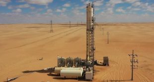 لقطة من الأعلى لمنشآت بئر مياه تابعة لمشروع النهر الصناعي في منطقة صحراوية شاسعة، تشمل برجاً معدنياً رأسياً وخزانات أفقية بيضاء ومعدات ضخ، ويمتد بجانبها صف من أعمدة بروج الكهرباء تحت سماء زرقاء بها غيوم متفرقة