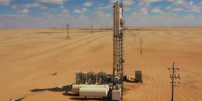 لقطة من الأعلى لمنشآت بئر مياه تابعة لمشروع النهر الصناعي في منطقة صحراوية شاسعة، تشمل برجاً معدنياً رأسياً وخزانات أفقية بيضاء ومعدات ضخ، ويمتد بجانبها صف من أعمدة بروج الكهرباء تحت سماء زرقاء بها غيوم متفرقة