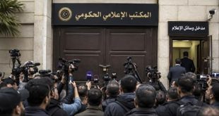تجمع لعدد من الصحفيين والمصورين يحملون الكاميرات والميكروفونات أمام البوابة الرئيسية المغلقة لـ "المكتب الإعلامي الحكومي" المقفلة بقفل حديدي، وإلى جانبها ممر فرعي مفتوح تعلوه لافتة مكتوب عليها "مدخل وسائل الإعلام