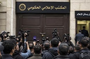 تجمع لعدد من الصحفيين والمصورين يحملون الكاميرات والميكروفونات أمام البوابة الرئيسية المغلقة لـ "المكتب الإعلامي الحكومي" المقفلة بقفل حديدي، وإلى جانبها ممر فرعي مفتوح تعلوه لافتة مكتوب عليها "مدخل وسائل الإعلام