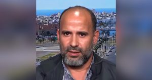 "لقطة مقربة (بورتريه) لرجل متوسط العمر ذي لحية خفيفة يتداخل فيها الشيب، يرتدي سترة داكنة فوق قميص مقلم، وتظهر خلفه شاشة تعرض خلفية لمدينة ساحلية مطلة على البحر تحت سماء صافية."