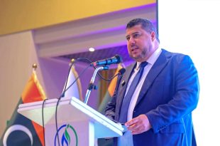 رئيس مجلس إدارة المؤسسة الوطنية للنفط مسعود سليمان يرتدي بدلة رسمية كحلية ويقف خلف منصة تتضمن شعار المؤسسة والعلم الليبي متحدثاً عبر الميكروفون في ورشة عمل حول تطوير صناعة الغاز الطبيعي