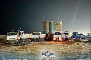 لقطة ليلية تظهر مجموعة من الشاحنات الكبيرة ومركبات النقل الثقيل متوقفة في موقع بناء وتجهيز ترابي، وفي الخلفية تظهر أبراج سكنية حديثة قيد الإنشاء مع رافعة برجية مضاءة.