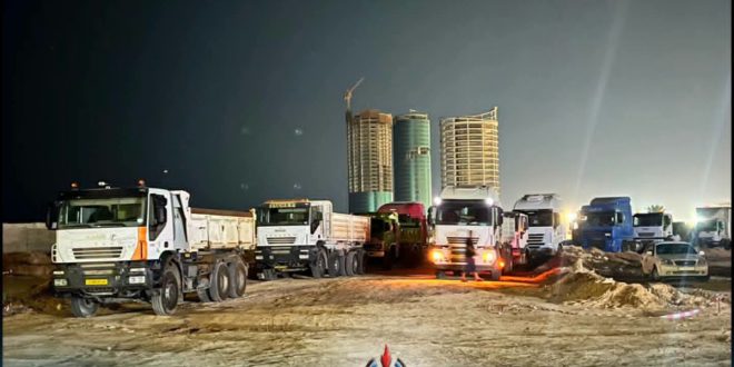 لقطة ليلية تظهر مجموعة من الشاحنات الكبيرة ومركبات النقل الثقيل متوقفة في موقع بناء وتجهيز ترابي، وفي الخلفية تظهر أبراج سكنية حديثة قيد الإنشاء مع رافعة برجية مضاءة.