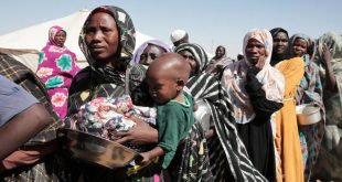 طابور طويل لنساء وأطفال نازحين في السودان يحملون أواني فارغة بانتظار المساعدات الغذائية داخل معسكر إيواء.