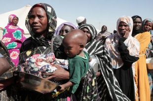 طابور طويل لنساء وأطفال نازحين في السودان يحملون أواني فارغة بانتظار المساعدات الغذائية داخل معسكر إيواء.