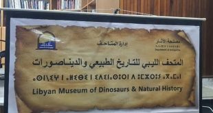 لوحة تعريفية تحمل اسم المتحف الليبي للتاريخ الطبيعي والديناصورات باللغتين العربية والإنجليزية، تتوسطها كتابة برموز قديمة، وتظهر عليها شعارات مصلحة الآثار وإدارة المتاحف، خلال فعالية أو عرض تعريفي داخل قاعة.
